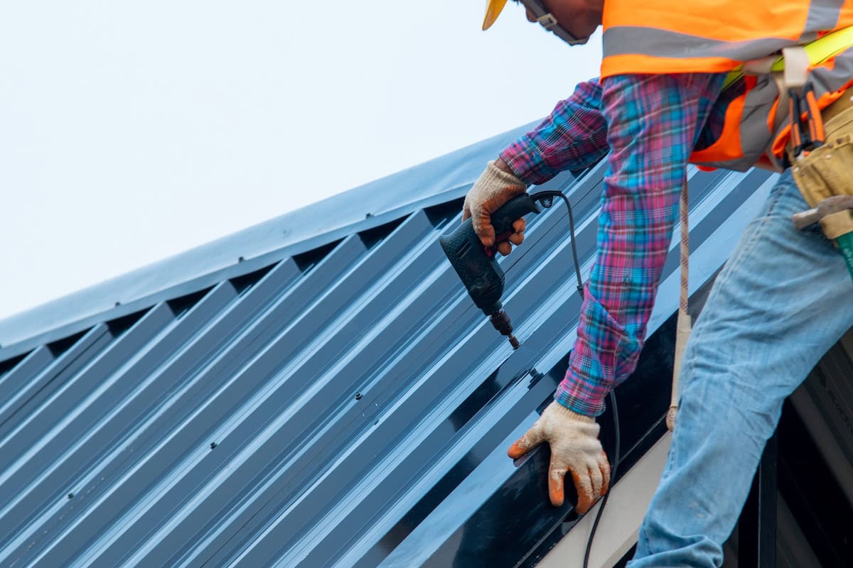 Worker in safety vest drills screws into a dark metal roof panel