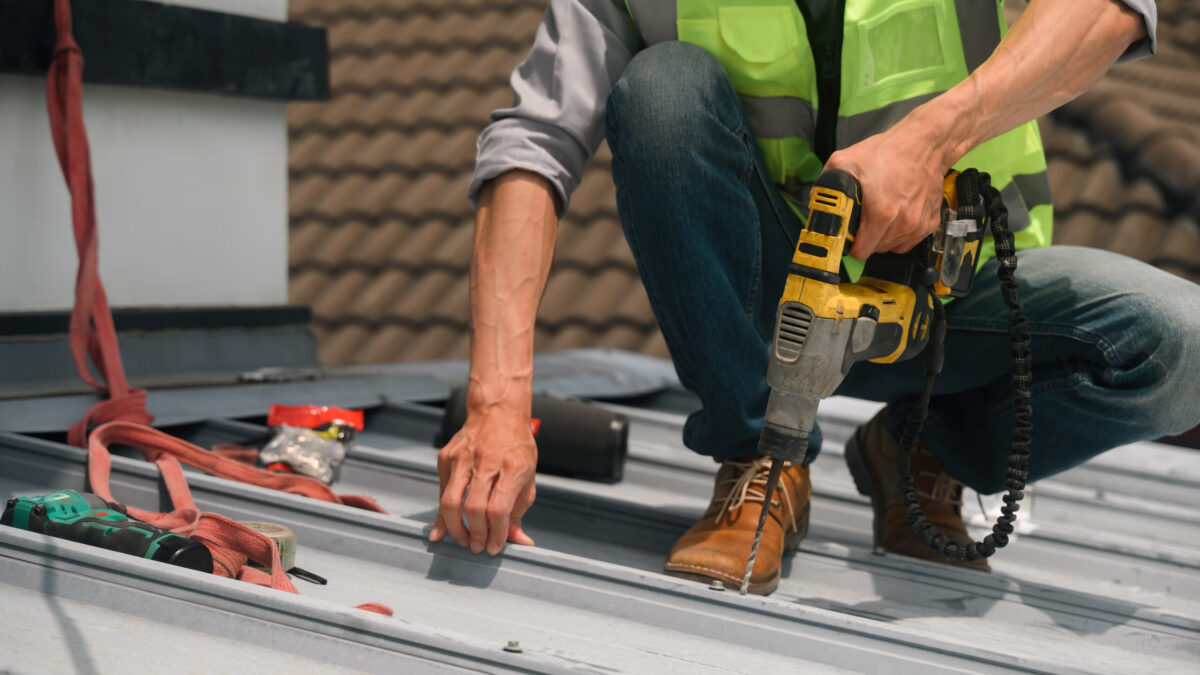 how long do metal roofs last Close up of a professional roofer in a safety vest drills into a metal roof panel during maintenance work, with tools and harness visible nearby.
